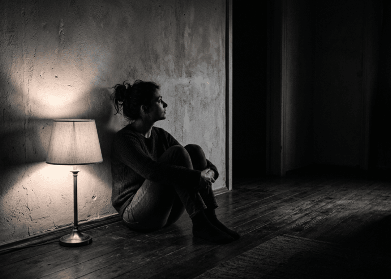 woman sitting against a wall in a dim room looking toward an open doorway, illustrating trauma and intuition